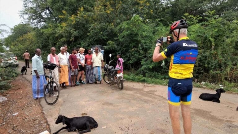 A group of people, some on bicycles, gathered on a dirt road in a rural area with lush greenery surrounding them. A cyclist in a bright jersey is taking a photo of the group.