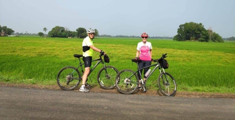 Two cyclists in colorful attire pause with their bikes on a rural road, surrounded by green fields and distant trees under a clear sky.