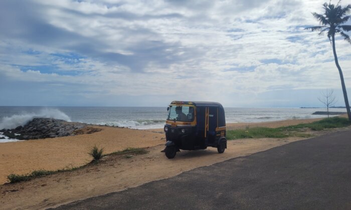 A black and yellow tuk tuk, a popular three-wheeled vehicle, parked on a sandy beach with a palm tree in the background. The image shows a coastal scene with waves crashing against the rocks and a cloudy sky overhead.