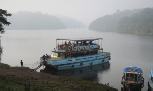 Scenic view of a large blue boat on a misty lake surrounded by lush green trees and hills in the distance. The boat appears to be a public transportation vessel used for sightseeing tours or commuting on the waterway.