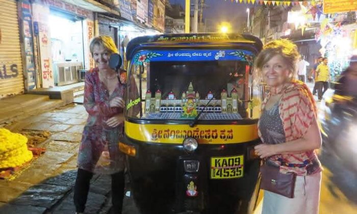 Two women posing beside a decorated auto rickshaw in a vibrant street setting, showcasing local culture and transportation.