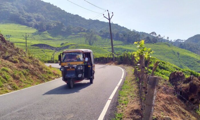 A three-wheeled tuk tuk vehicle driving along a winding road through a scenic countryside landscape with lush green hills, tea plantations, and power lines in the background.