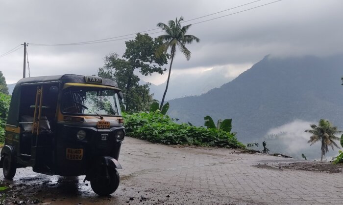 A black and yellow auto-rickshaw, commonly known as a tuk-tuk, parked on a wet, paved road surrounded by lush green vegetation and mountains in the background. The image suggests a scenic, rainy landscape in Kerala, India, where tuk-tuks are a popular mode of transportation.