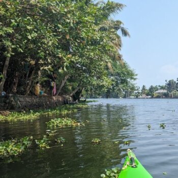 A panoramic view of a serene lake surrounded by lush green hills, in Alleppey, with the clear blue sky enhancing the natural beauty of the landscape.