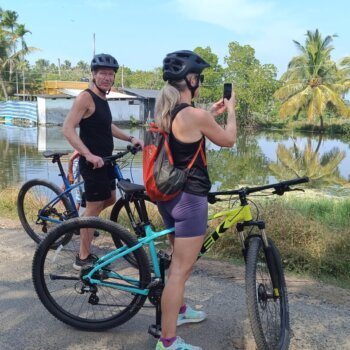 Two cyclists wearing helmets stop to take photos of a scenic lake surrounded by lush palm trees, on a sunny day.