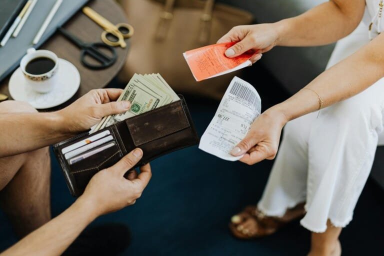 A man passes cash to another while both sit on a couch.