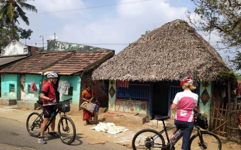 Chidambaram to Thanjavur Cyclists stopping in front of traditional thatched-roof houses in a rural village setting, interacting with local residents.